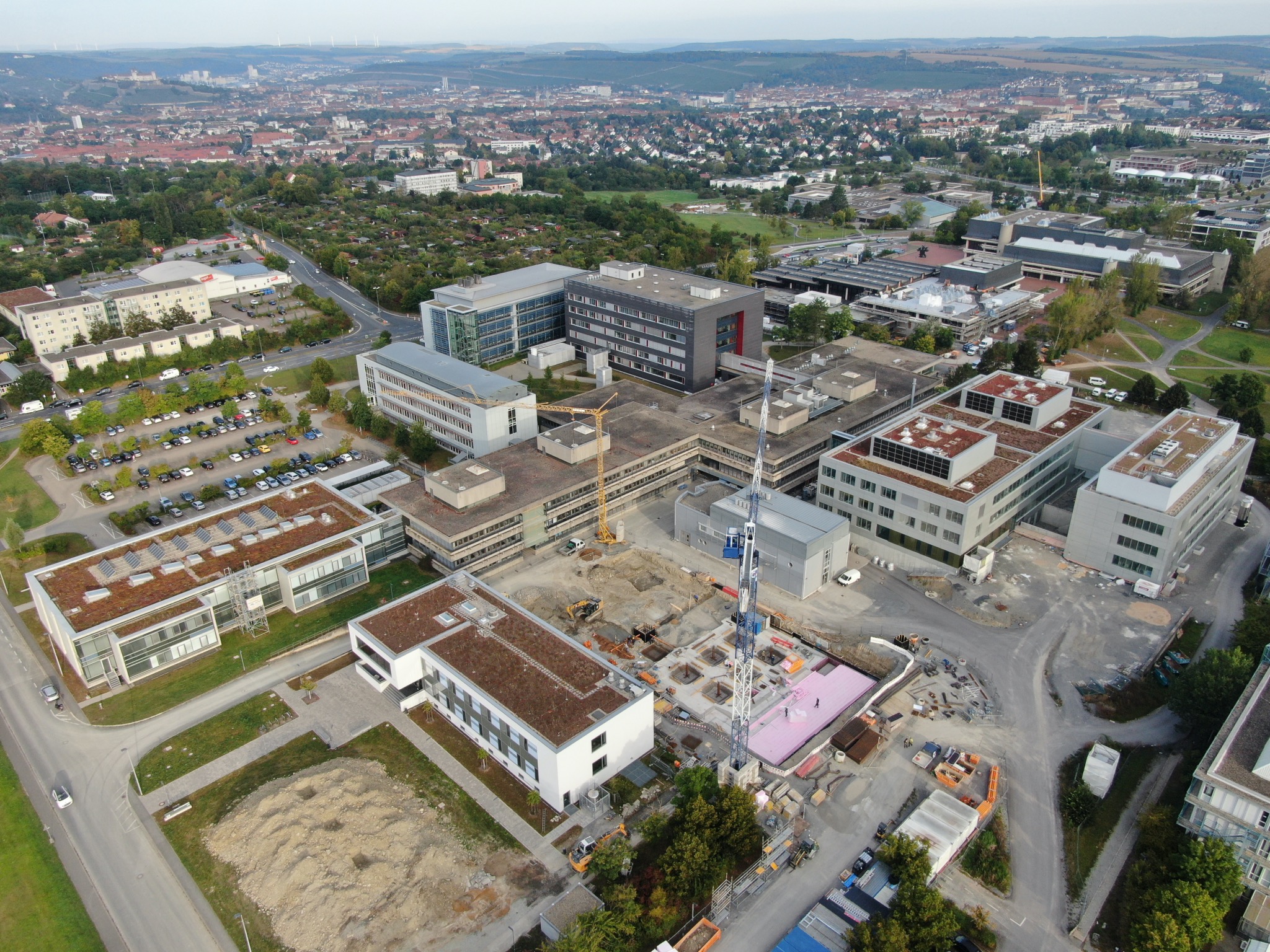 Übersicht über die Gebäude der Fakultät für Chemie und Pharmazie am Hubland. In der Mitte ist der Zentralbau zu sehen, der noch saniert werden muss. In dem Bereich zwischen den beiden Baukränen findet das neue Praktikumsgebäude Platz; auf der Wiese mit dem Erdhaufen am linken unteren Bildrand entsteht das Center of Polyerms for Life. Foto: Staatliches Bauamt Würzburg