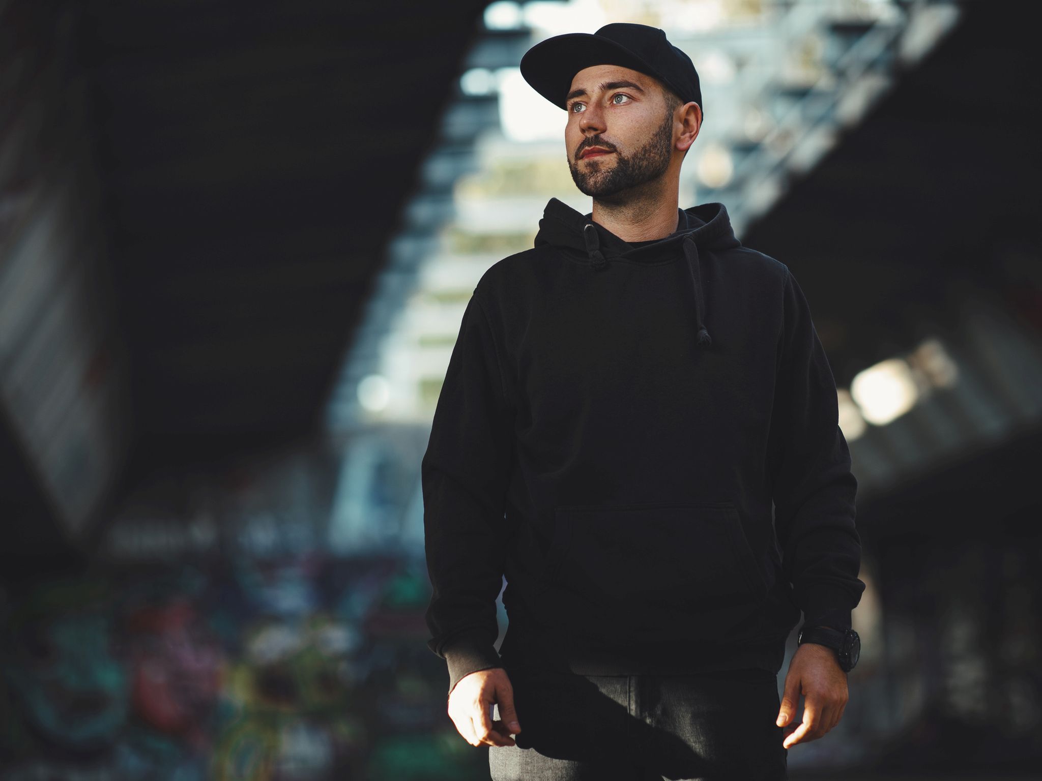 Portrait of a young bearded man with a black hat ain black hoodie in the city