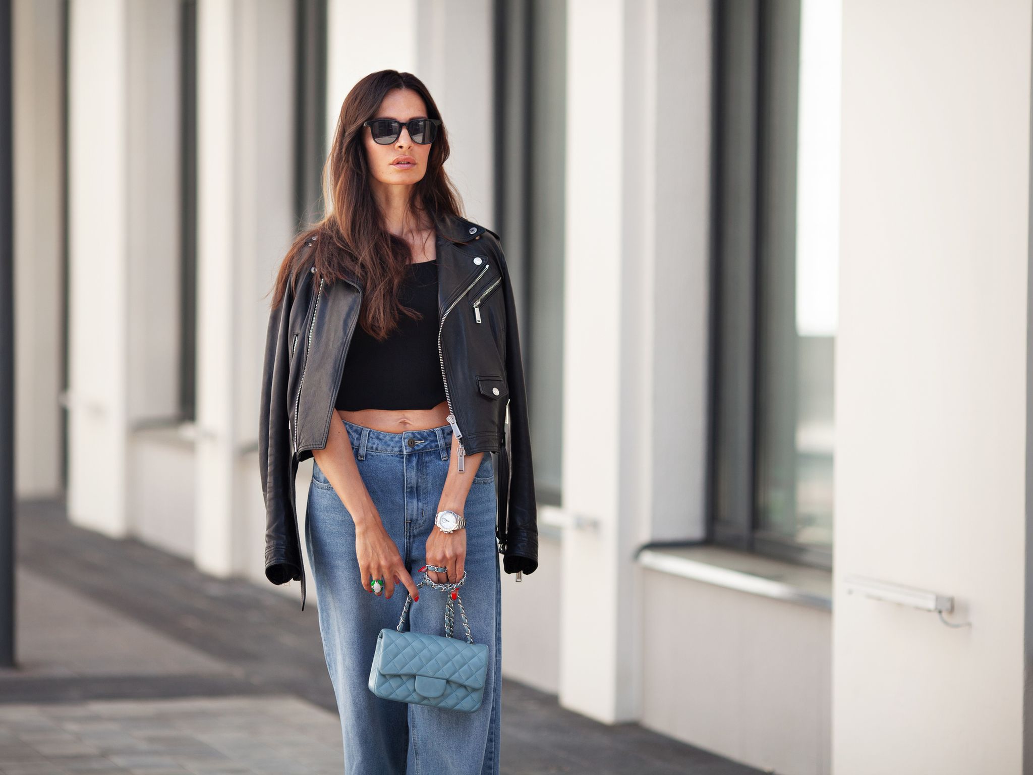 Portrait of gorgeous brunette woman standing city street. Fashion model wears black leather jacket, jeans, short top, small handbag on chain. Street style
